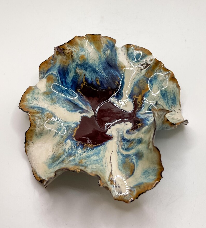 May include: A handcrafted ceramic bowl with an organic shape. The bowl has a mix of colours: deep burgundy, blues, cream, and brown. The edges are slightly raised, and the surface has a glossy finish. The bowl is approximately 25 cm in diameter.