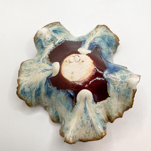 May include: A handmade ceramic bowl with an unusual, natural form. It has a deep burgundy centre, framed by a white and blue glazed rim. The edges have a textured, earthy look, enhancing its artistic quality. The bowl is likely for decorative use.