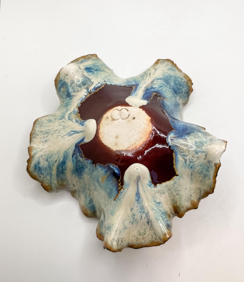 May include: A handmade ceramic bowl with an unusual, natural form. It has a deep burgundy centre, framed by a white and blue glazed rim. The edges have a textured, earthy look, enhancing its artistic quality. The bowl is likely for decorative use.