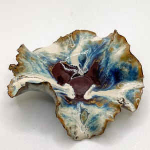 May include: A ceramic bowl with an unusual, natural form. The bowl has a deep burgundy centre, with blue, cream, and gold tones. The rim is textured and wavy, enhancing its artistic quality. The bowl's design is reminiscent of a flower.