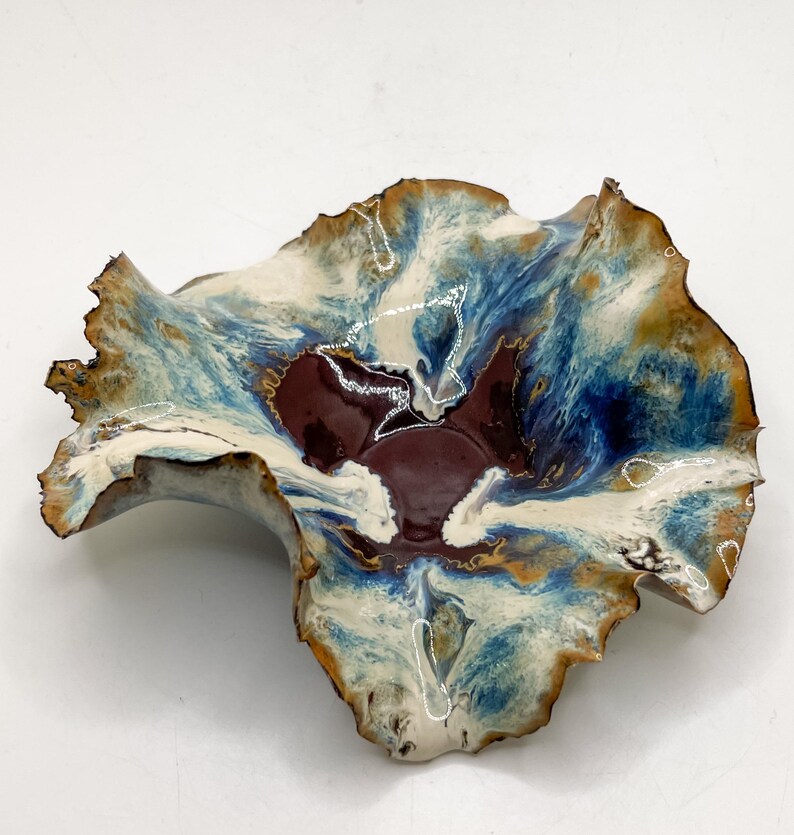 May include: A ceramic bowl with an unusual, natural form. The bowl has a deep burgundy centre, with blue, cream, and gold tones. The rim is textured and wavy, enhancing its artistic quality. The bowl's design is reminiscent of a flower.