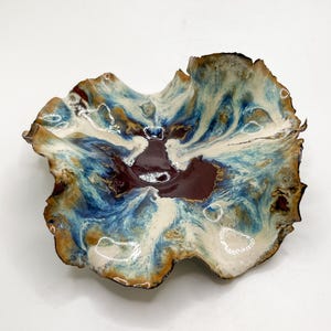 May include: A handmade ceramic bowl with an unusual, natural form. The bowl has a rich colour scheme of deep burgundy, creamy white, and shades of blue and brown. The edges are textured, enhancing its artistic quality. This decorative bowl is suitable for serving or display.