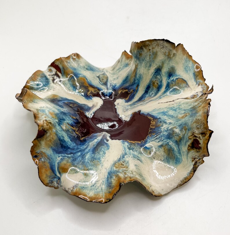 May include: A handmade ceramic bowl with an unusual, natural form. The bowl has a rich colour scheme of deep burgundy, creamy white, and shades of blue and brown. The edges are textured, enhancing its artistic quality. This decorative bowl is suitable for serving or display.