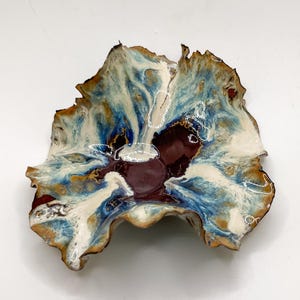 May include: A handmade ceramic bowl with an irregular, natural form. The bowl showcases a mix of colours: deep burgundy, creamy white, and blue and brown tones. Fine white lines decorate the surface, enhancing the artistic design of the bowl.