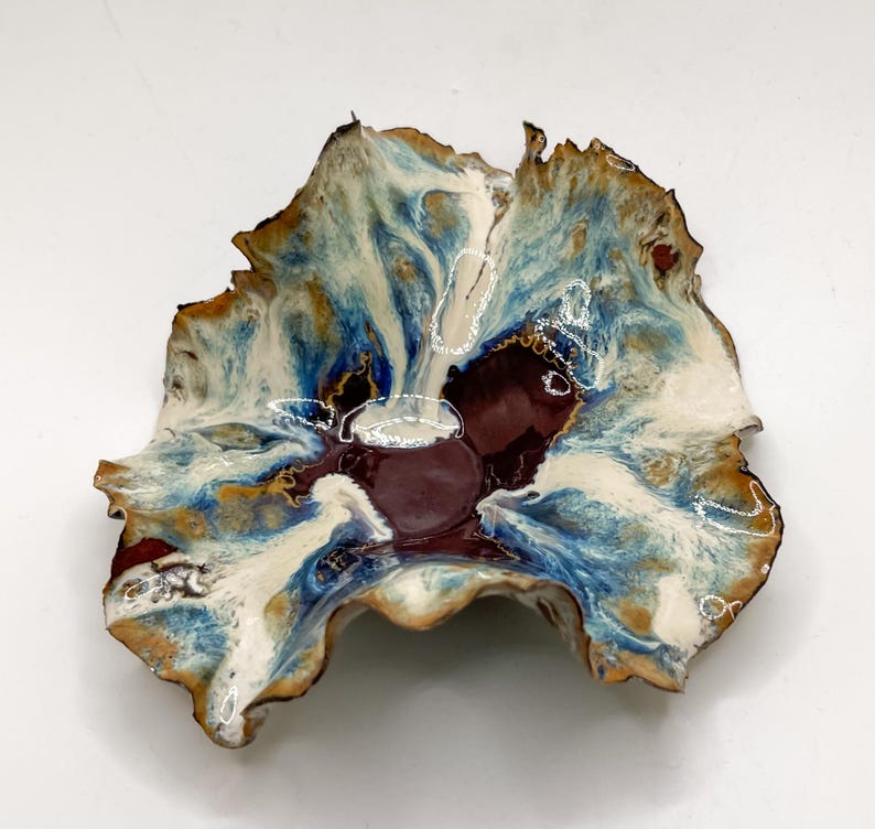 May include: A handmade ceramic bowl with an irregular, natural form. The bowl showcases a mix of colours: deep burgundy, creamy white, and blue and brown tones. Fine white lines decorate the surface, enhancing the artistic design of the bowl.