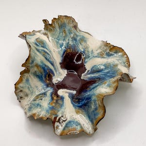 May include: A handmade ceramic bowl with an unusual, natural form. The bowl displays a mix of colours, including deep burgundy, white, and shades of blue and brown. The edges have a textured, irregular look, enhancing its artistic charm.