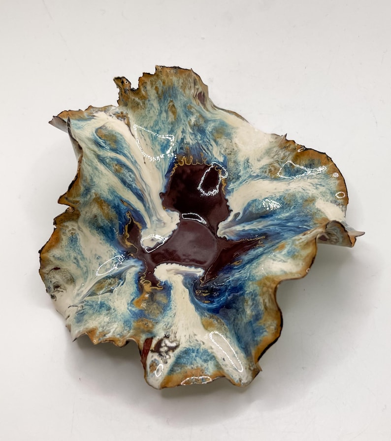 May include: A handmade ceramic bowl with an unusual, natural form. The bowl displays a mix of colours, including deep burgundy, white, and shades of blue and brown. The edges have a textured, irregular look, enhancing its artistic charm.
