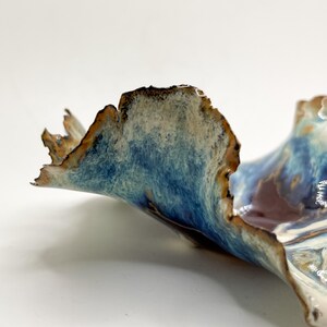 May include: A ceramic bowl with an unusual, natural form. It has a blue, brown, and white glaze, giving a textured, wave-like look. The edges are rough, enhancing its artistic quality. A decorative piece for the home.