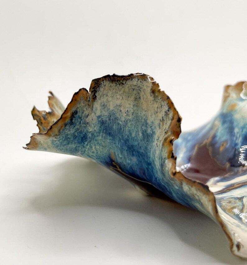 May include: A ceramic bowl with an unusual, natural form. It has a blue, brown, and white glaze, giving a textured, wave-like look. The edges are rough, enhancing its artistic quality. A decorative piece for the home.