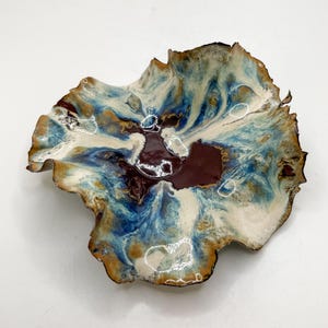 May include: A decorative ceramic bowl with an unusual, organic form. The bowl has a swirling pattern of blue, white, brown, and maroon colours. The rim has a dark brown edge. This handcrafted bowl is ideal for serving or display.