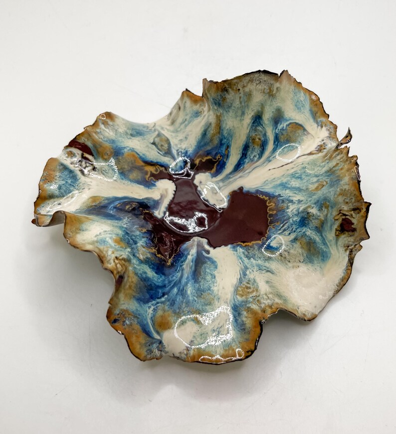 May include: A decorative ceramic bowl with an unusual, organic form. The bowl has a swirling pattern of blue, white, brown, and maroon colours. The rim has a dark brown edge. This handcrafted bowl is ideal for serving or display.