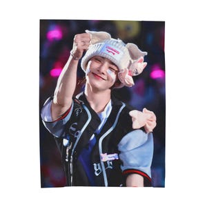 May include: A person wearing a white knit hat with attached ears and a pink flower. The person is wearing a black and blue baseball-style jacket with white accents. The background is blurred with pink and purple lights.