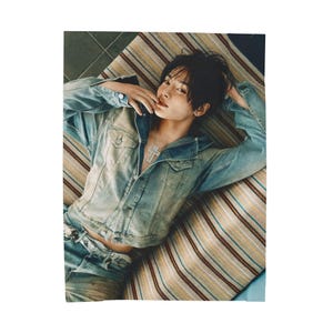 May include: A light blue denim jacket and matching jeans. The jacket has buttoned pockets and a collar. A silver cross pendant hangs from a chain. The person is lying on a striped cushion.