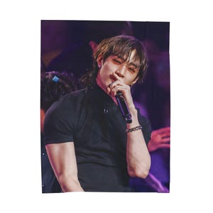 May include: A black short-sleeved shirt with a high collar. The person in the image is holding a microphone. The background is a mix of purple and dark colors. The person is wearing a bracelet on their wrist.