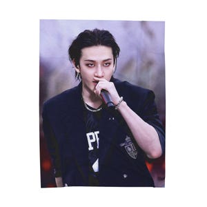 May include: A rectangular wall tapestry featuring a person holding a microphone. The person is wearing a black blazer over a black shirt with white lettering. The background is a blurred mix of purple and red.