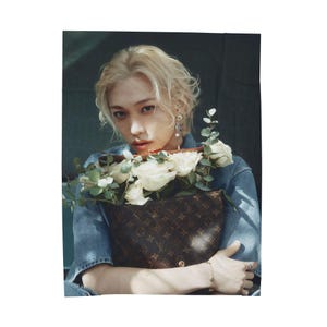 May include: A person holding a brown handbag filled with white roses and greenery. The handbag features a repeating logo pattern. The person is wearing a denim shirt and has blonde hair.