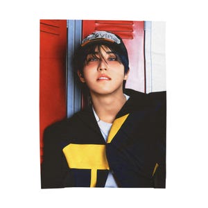 May include: A person wearing a black jacket with yellow accents and a baseball cap with the word "Vying" on it. The person is leaning against a red locker.