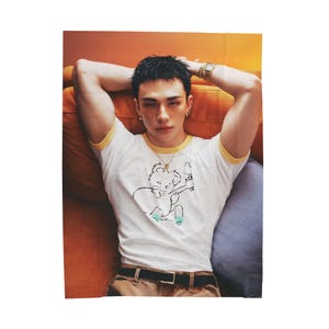 May include: A person wearing a white t-shirt with a yellow trim and a koala graphic. The person is wearing a gold necklace, a watch, and earrings. They are lying on a brown and blue surface.