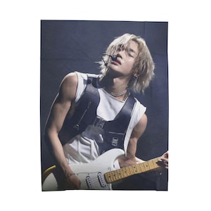 May include: A white electric guitar with a maple neck is held by a person wearing a white sleeveless shirt and a black vest. The person has blonde hair and is wearing a silver necklace and bracelet. The background is dark.