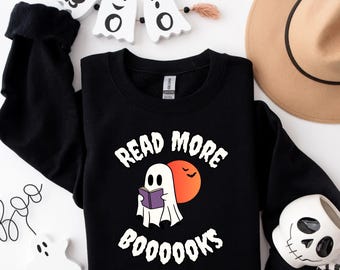 May include: Black sweatshirt with a Halloween-themed graphic. The design features a ghost reading a book with the text "READ MORE BOOOOOKS". An orange moon and two bats are also included in the design. Additional Halloween decorations are in the background.