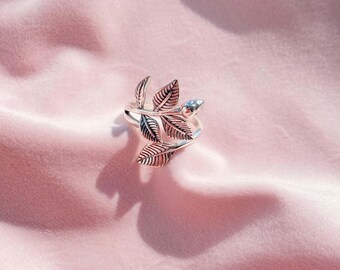 Könnte beinhalten: Ein silberfarbener Ring mit Blattdesign. Der Ring hat mehrere detaillierte Blätter, die sich um den Finger wickeln. Ein kleiner, hellfarbiger Stein ist am Ende des Rings eingesetzt. Der Ring wird auf einer rosa, samtigen Oberfläche präsentiert.