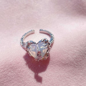 May include: A silver ring with a white heart-shaped stone surrounded by smaller clear stones. The ring is on a pink background.