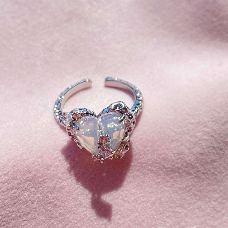 May include: A silver ring with a white heart-shaped stone surrounded by smaller clear stones. The ring is on a pink background.