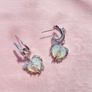 May include: A pair of silver earrings with a heart-shaped charm. The charms are adorned with a pale green stone and clear rhinestones. The earrings have a hoop design.