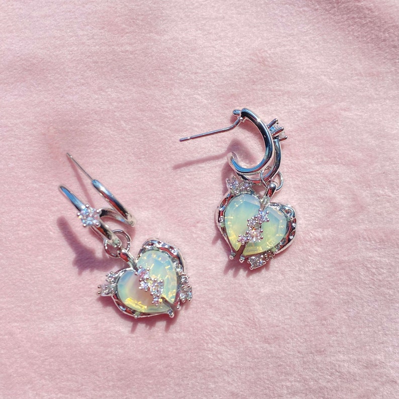 May include: A pair of silver earrings with a heart-shaped charm. The charms are adorned with a pale green stone and clear rhinestones. The earrings have a hoop design.