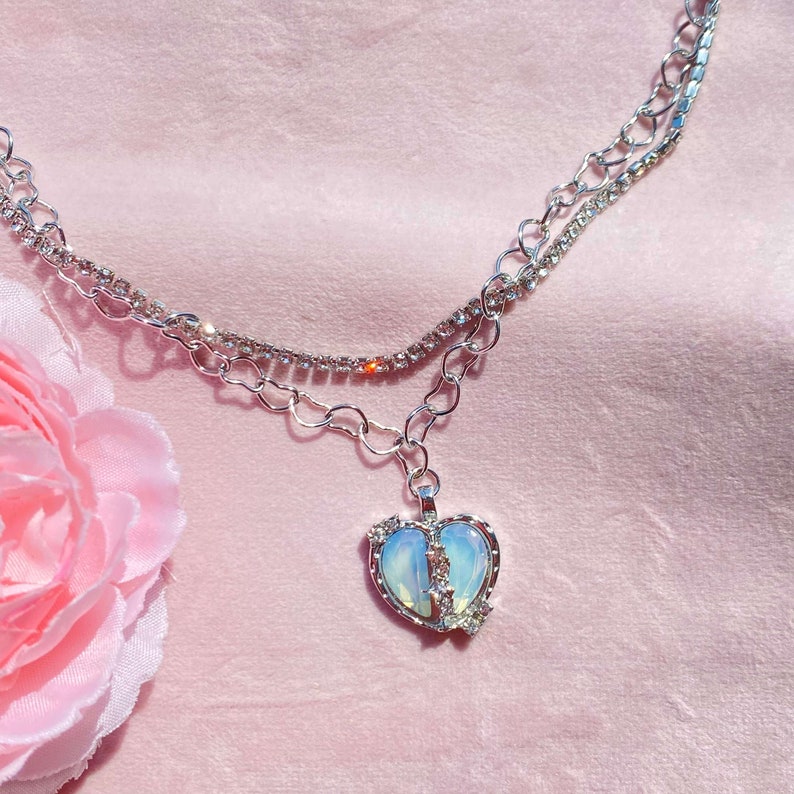 May include: A silver chain necklace with a broken heart pendant. The heart is made of iridescent glass and has rhinestones around the edges. The chain is layered with a smaller chain of rhinestones.