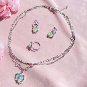 May include: A silver necklace with a heart pendant, a matching ring, and a pair of heart-shaped earrings. The jewelry features iridescent stones and a delicate chain.