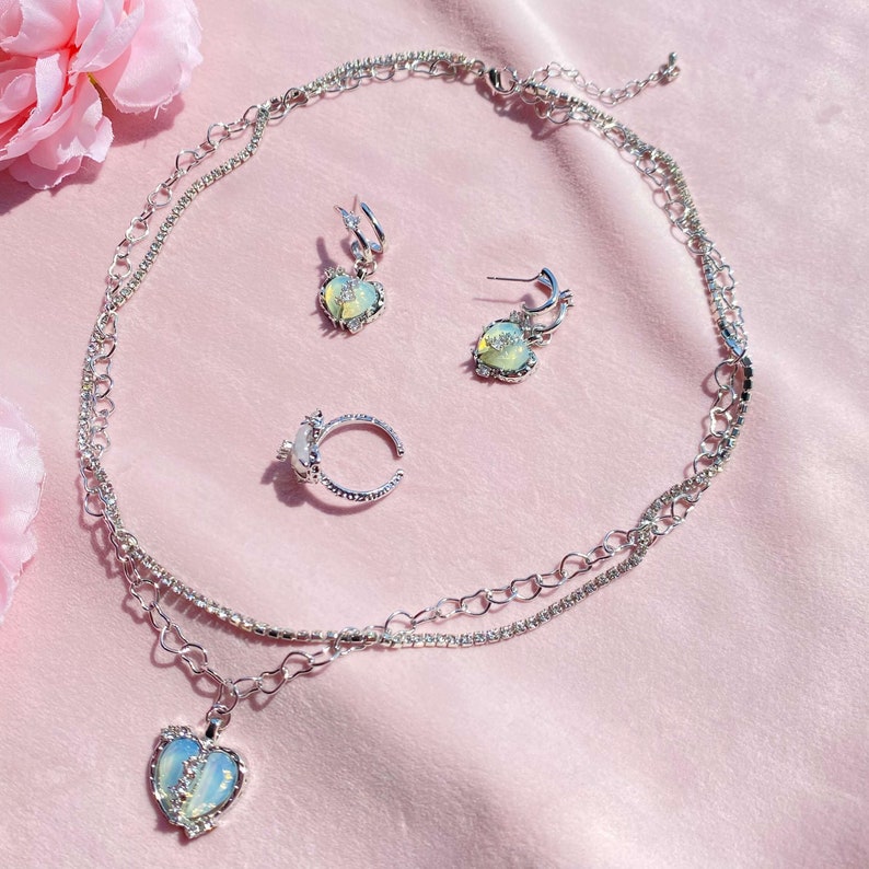 May include: A silver necklace with a heart pendant, a matching ring, and a pair of heart-shaped earrings. The jewelry features iridescent stones and a delicate chain.