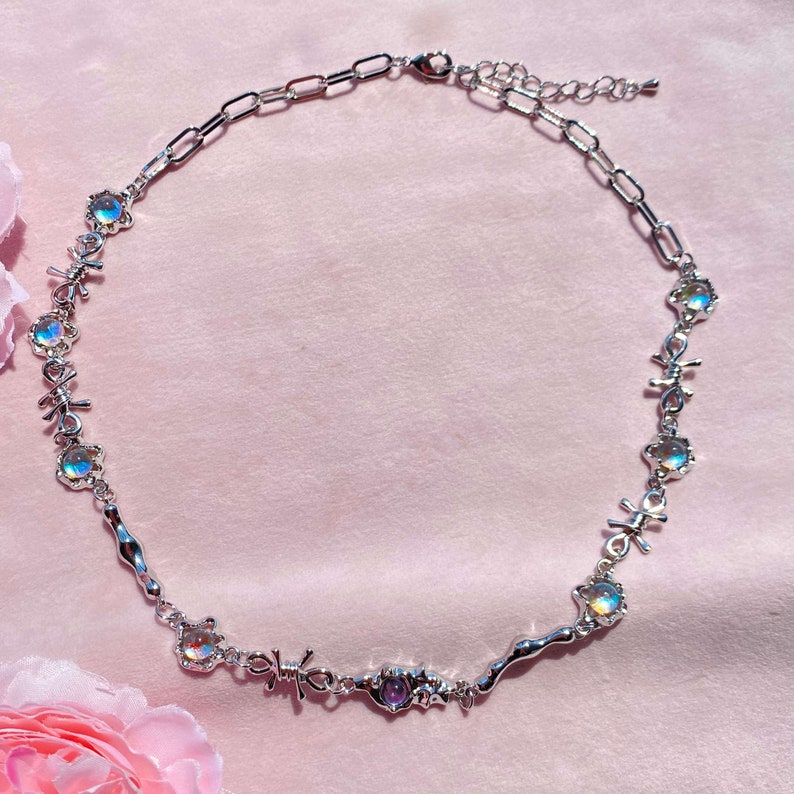 May include: A silver necklace with iridescent stones and a barbed wire design. The necklace has a lobster clasp closure and an extender chain.