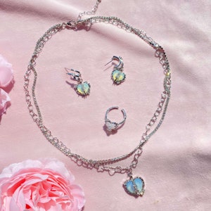May include: A silver necklace with a heart pendant, a pair of silver earrings with heart pendants, and a silver ring with a heart pendant. All of the pendants have iridescent stones.