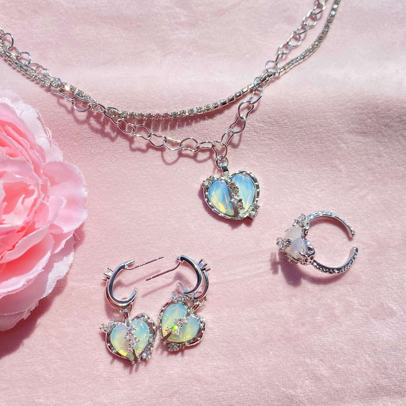 May include: A set of silver jewelry featuring a broken heart pendant necklace, a ring, and a pair of earrings. Each piece has a clear iridescent stone in the center and is adorned with small sparkling rhinestones.