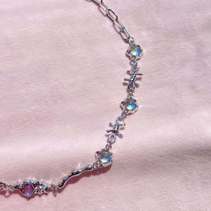 May include: A silver necklace with iridescent stones and barbed wire accents. The necklace is on a pink background.
