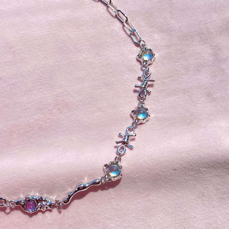 May include: A silver necklace with iridescent stones and barbed wire accents. The necklace is on a pink background.