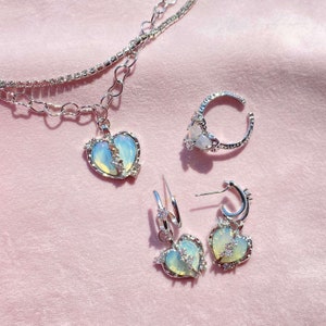 May include: A set of silver jewelry featuring a broken heart design. The set includes a necklace, a ring, and a pair of earrings. The heart pendants are made of iridescent opal-like glass with silver accents.