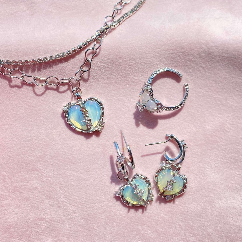 May include: A set of silver jewelry featuring a broken heart design. The set includes a necklace, a ring, and a pair of earrings. The heart pendants are made of iridescent opal-like glass with silver accents.
