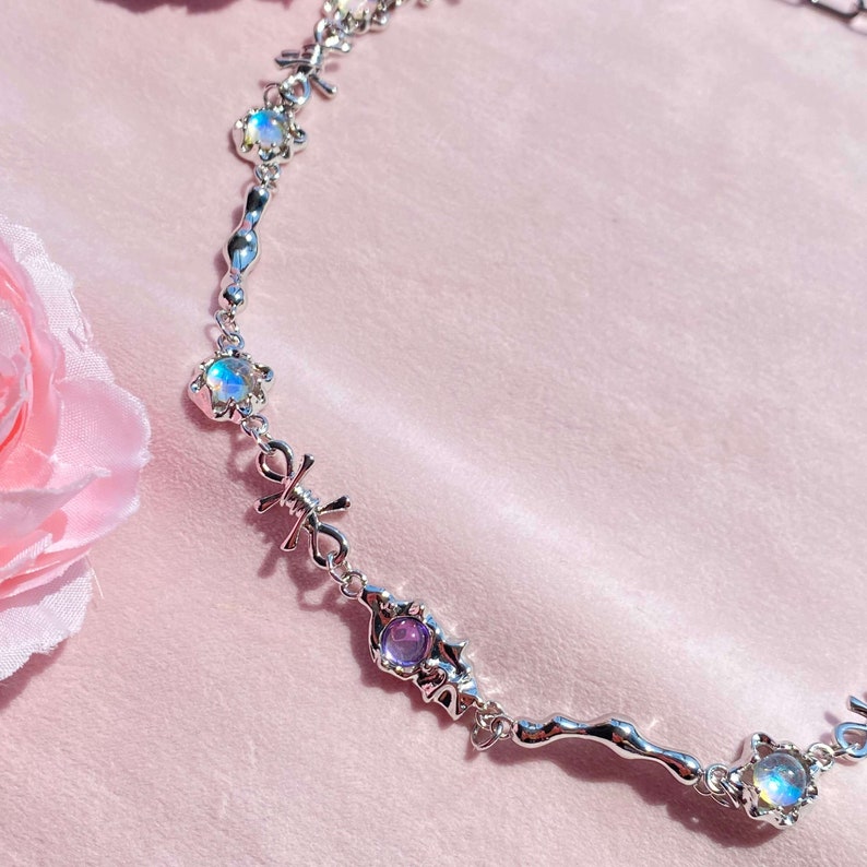 May include: A silver chain necklace with iridescent and purple gemstones. The chain features a repeating pattern of intertwined X shapes.