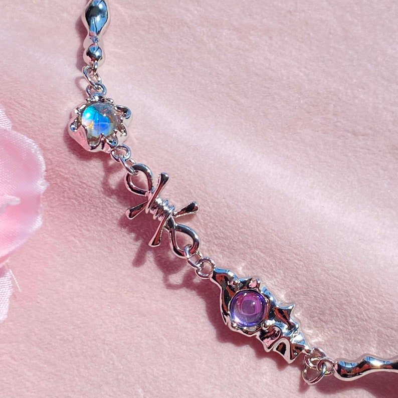 May include: A silver chain necklace with two iridescent gemstones and a barbed wire design. The necklace features a silver chain with a small, iridescent gemstone in a flower-shaped setting, followed by a barbed wire design, and then a larger, iridescent gemstone in a more intricate setting.