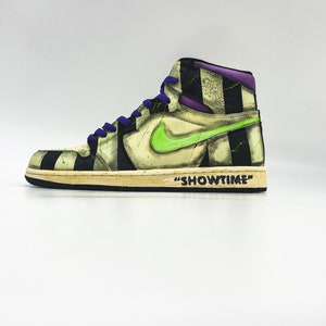 Puede incluir: Un par de zapatillas de baloncesto de caña alta con un patrón de rayas blancas y negras, detalles verdes y un swoosh morado. El texto "SHOWTIME" está escrito en la suela del zapato.