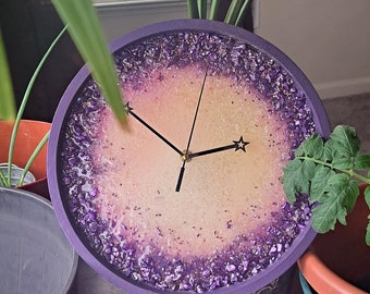 May include: A purple resin clock with a white face and gold hands. The clock is decorated with purple glitter and small purple stones around the edge.