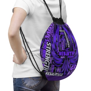 Peut inclure: Un sac à cordon de serrage violet avec les mots "Force", "Renaissance" et "Résilience" en blanc et noir. Le sac a un cordon noir et est porté sur l'épaule.