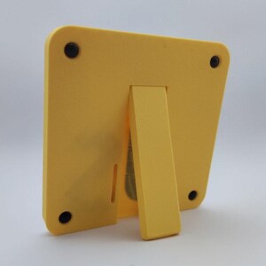 Pode incluir: Uma moldura amarela com um suporte embutido. A moldura tem cantos arredondados e quatro p&eacute;s de borracha pretos. O suporte &eacute; de cor amarela combinando e &eacute; fixado na parte de tr&aacute;s da moldura. A moldura foi projetada para ficar em p&eacute; em uma superf&iacute;cie plana.