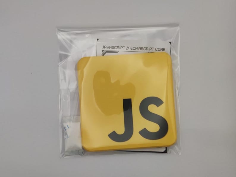 Pode incluir: Um objeto quadrado amarelo com as letras pretas "JS", embalado em um saco pl&aacute;stico transparente. O texto "JAVASCRIPT // ECMASCRIPT CORE" &eacute; vis&iacute;vel em um pequeno cart&atilde;o. O item parece ser um adesivo ou produto similar.