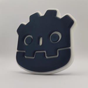 May include: A 3D printed blue and white robot head with a simple design. The robot has two eyes and a mouth.