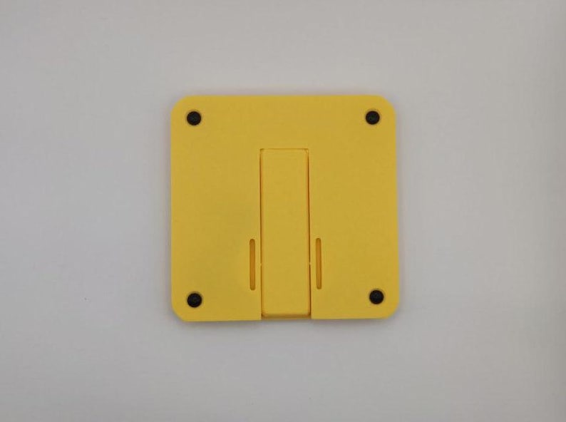 Pode incluir: Um suporte de telefone amarelo com uma base retangular e um suporte dobr&aacute;vel. O suporte tem quatro p&eacute;s circulares pretos e duas ranhuras para segurar um telefone. O suporte foi projetado para apoiar um telefone para uso m&atilde;os-livres.
