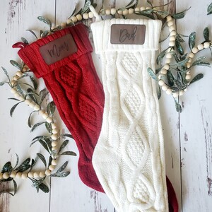 May include: Two knitted Christmas stockings with leather name tags. The stocking on the left is red and says "Mom". The stocking on the right is white and says "Dad".