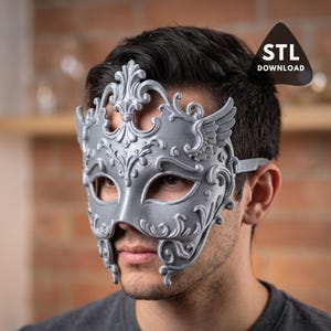 May include: A silver-colored masquerade mask with ornate, swirling details and wing accents. The mask covers the eyes and nose, with a strap to secure it. The image includes the text "STL DOWNLOAD".
